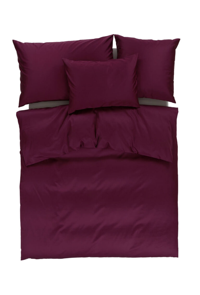 Ein ordentlich gemachtes Bett mit einer weinroten Bettdecke, zwei passenden Kissen am Kopfende und einem kleineren Kissen, das waagerecht vor ihnen liegt, alles vor einem weißen Hintergrund.