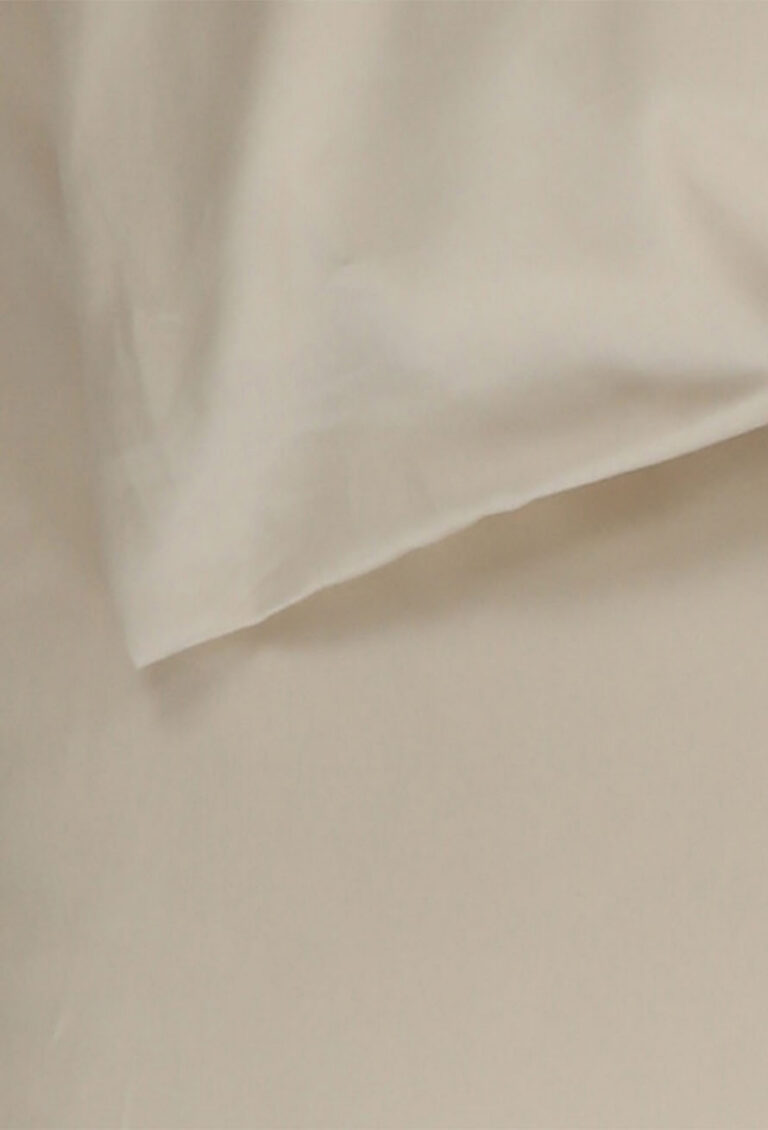 Eine Nahaufnahme eines beigen Kissens, das auf einem passenden beigen Bettlaken liegt, das glatte, weiche Gewebe mit minimaler Textur zeigt.