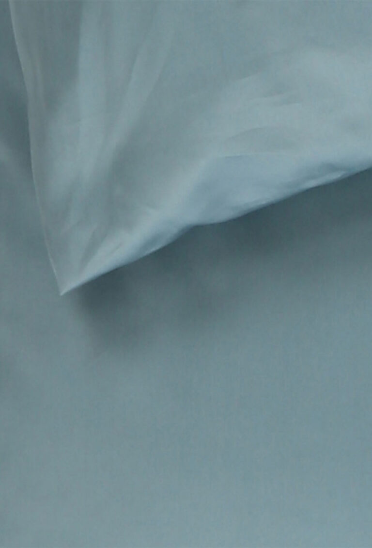 Eine Nahaufnahme eines hellblauen Kissens auf einem passenden hellblauen Bettlaken, das glatten, leicht zerknitterten Stoff zeigt.