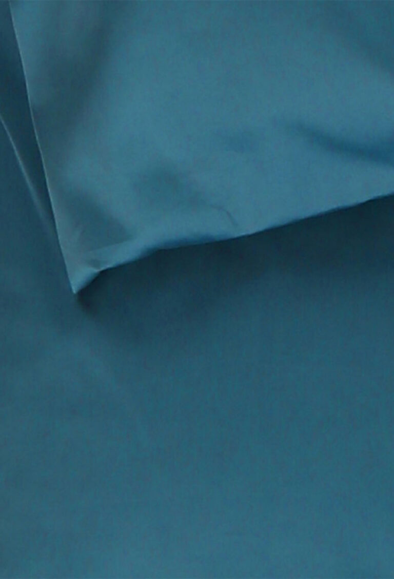 Nahaufnahme eines blauen Bettlakens und eines dazu passenden Kopfkissenbezugs, beide aus glattem Stoff. Der Kopfkissenbezug ist schräg in der oberen linken Ecke des Bildes positioniert.