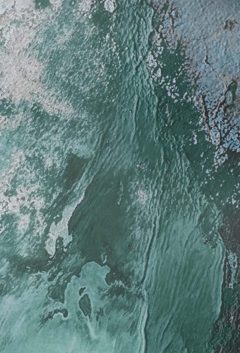 Luftaufnahme einer grün und blau strukturierten Oberfläche, die an Wasser mit Wellen und Flecken aus helleren blauen und weißen Bereichen, möglicherweise Eis oder Schaum, im oberen und rechten Teil des Bildes erinnert.