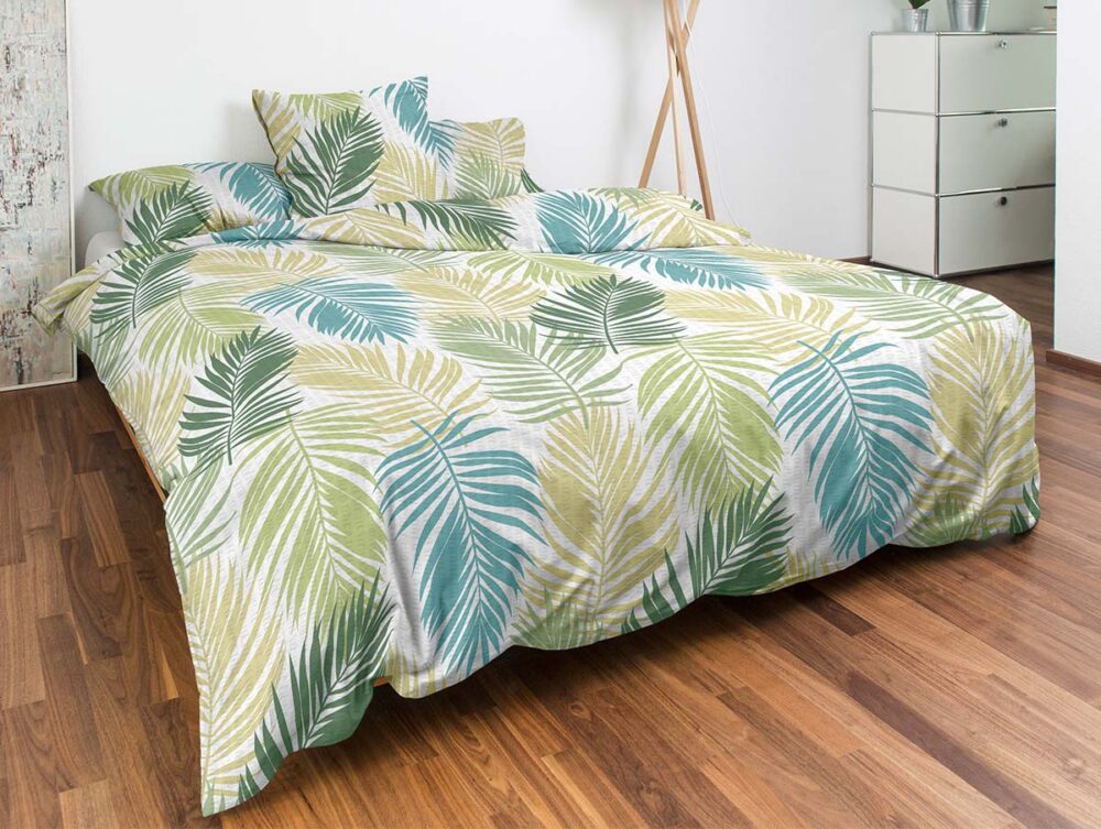Ein ordentlich gemachtes Bett mit Bettwäsche mit grünem und gelbem Palmenblattmuster steht auf einem Holzboden. Dazu gibt es passende Kissen, eine moderne Lampe und eine weiße Kommode im Hintergrund.