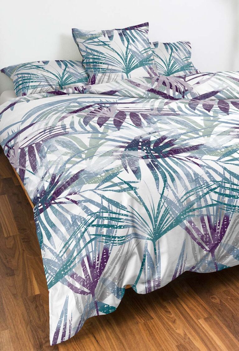 Ein Bett mit einer weißen Bettdecke und Kissen mit einem tropischen Blattmuster in Teal-, Grau- und Violetttönen in einem Raum mit Holzfußboden und einer schlichten weißen Wand.