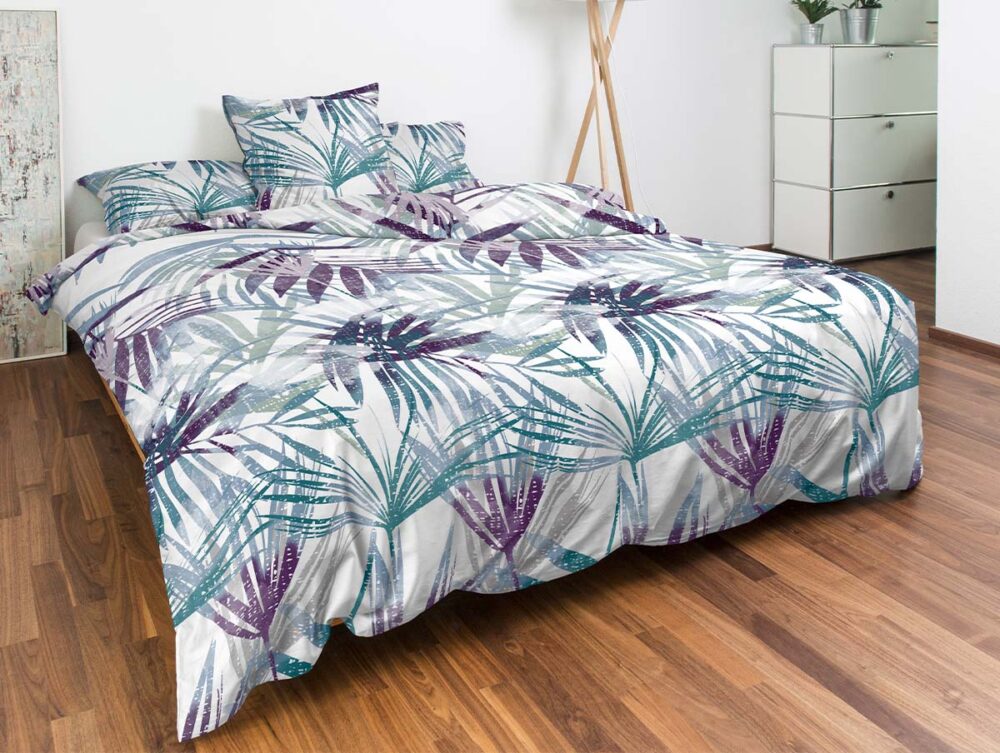 Ein ordentlich gemachtes Bett mit weißer, blaugrüner und violetter, blattgemusterter Bettwäsche, passenden Kissen und einem Holzrahmen in einem Zimmer mit Holzböden, einer Dreifußlampe, weißen Schubladen und einer Topfpflanze.