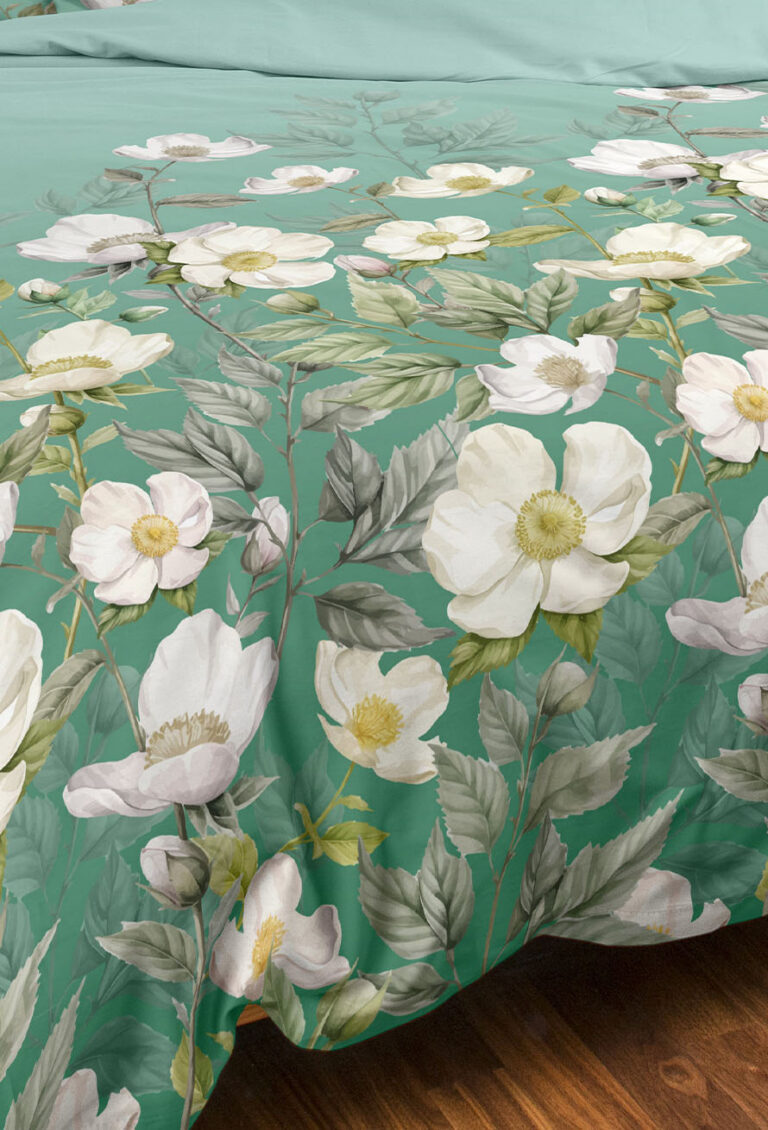 Nahaufnahme einer grünen Tagesdecke, die mit großen weißen Blumen und blattartigen Zweigen verziert ist und ein detailliertes botanisches Muster auf einem Holzboden zeigt.