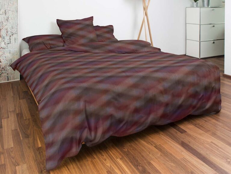 Ein ordentlich gemachtes Bett mit einer dunklen, bunt gemusterten Bettdecke und Kissen, in einem modernen Schlafzimmer mit Holzfußboden, weißen Wänden, einer Kommode und einer Stehlampe.