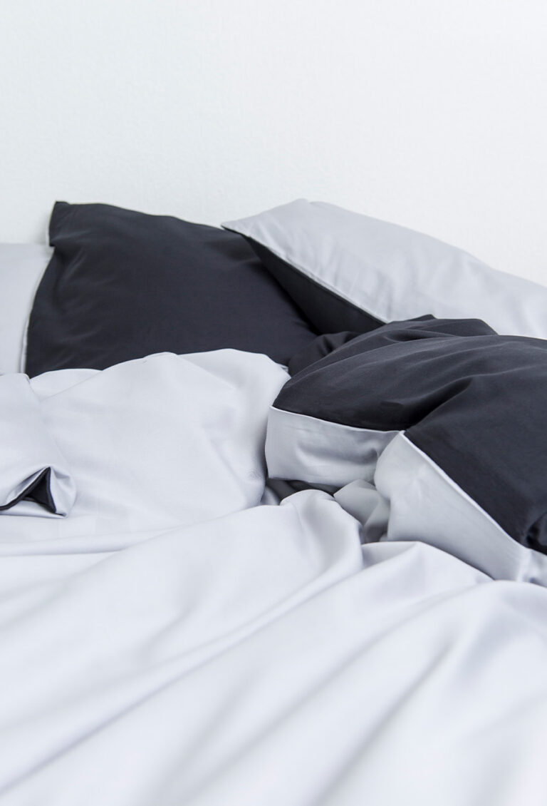 Ein ordentlich gemachtes Bett mit Bettwäsche Nobilium Mia silber/anthrazit, zwei schwarz-grauen Kissen und einer weißen Wand im Hintergrund.