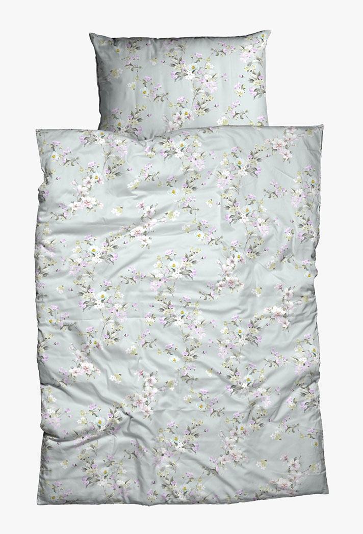 Ein Bett mit einem passenden Kissen und einer Bettdecke mit hellgrauem Hintergrund und einem zarten Blumenmuster mit kleinen rosa, weißen und gelben Blumen und grünen Zweigen.