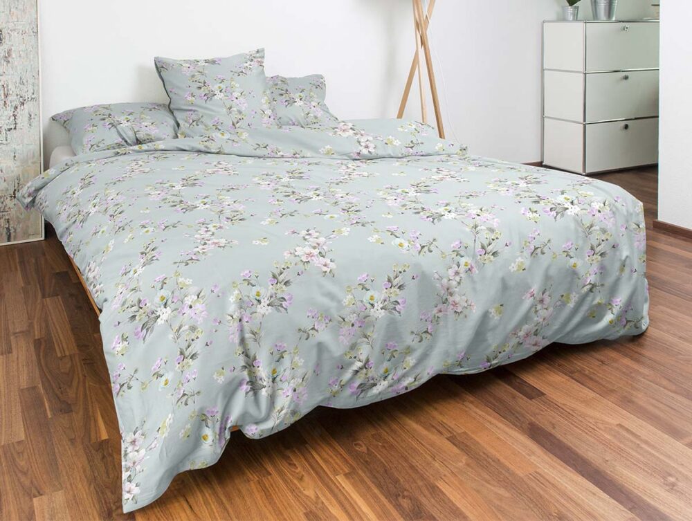Ein ordentlich gemachtes Bett mit hellblauer Bettwäsche mit zartem Blumenmuster in einem modernen Schlafzimmer mit Holzboden, einer weißen Kommode und einer Stehlampe im Hintergrund.