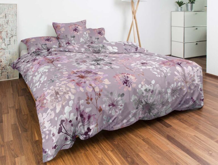 Ein ordentlich gemachtes Bett mit einer lila und rosa geblümten Bettdecke und Kissen steht auf einem Holzboden. In der Nähe befinden sich eine Stehlampe, Kunstwerke, eine weiße Kommode und eine kleine Pflanze in einem modernen, hellen Schlafzimmer.