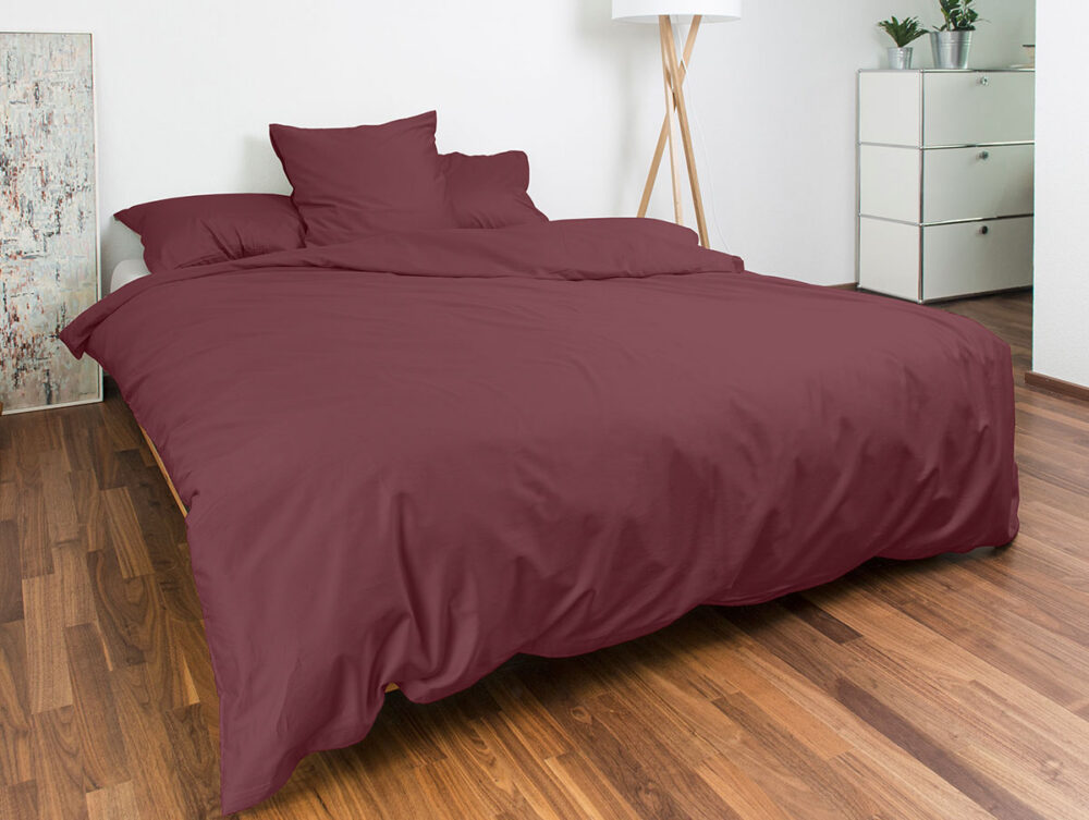 Ein ordentlich gemachtes Bett mit burgunderrotem Bettzeug und drei Kissen steht in einem modernen Schlafzimmer mit Holzfußboden, einer weißen Kommode mit Topfpflanzen, einer Stehlampe und einem abstrakten Wandkunstwerk.