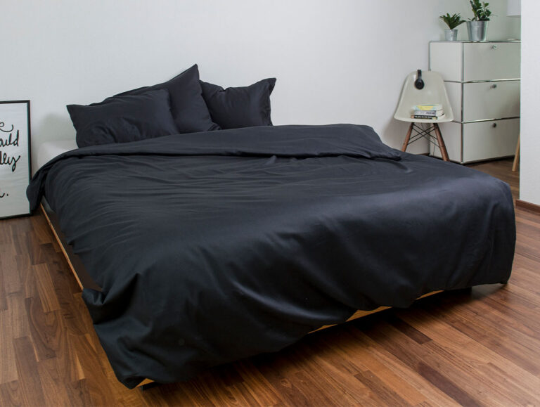 Ein ordentlich gemachtes Bett mit schwarzem Bettzeug und Kissen steht auf einem Holzboden in einem minimalistischen Schlafzimmer. Im Hintergrund stehen ein weißer Stuhl, eine kleine Kommode mit Pflanzen und ein gerahmtes Bild.