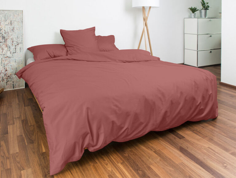 Ein ordentlich gemachtes Bett mit gedämpfter rosa Bettwäsche in einem modernen Schlafzimmer mit Holzfußboden, einer Stehlampe, einer weißen Kommode mit Pflanzen und einem großen angelehnten Kunstwerk in der Ecke.