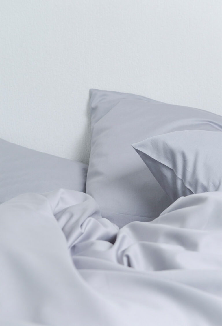 Zwei hellgraue Kissen und eine dazu passende zerknitterte Bettdecke sind auf einem Bett vor einer weißen Wand angeordnet und schaffen einen weichen und minimalistischen Look.