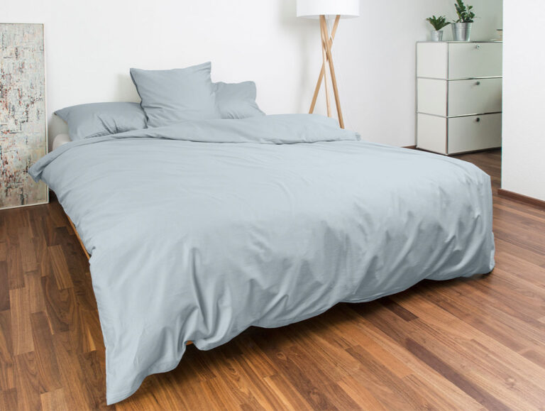 Ein ordentlich gemachtes Bett mit hellgrauer Bettwäsche in einem modernen Schlafzimmer mit Holzboden, einer weißen Kommode, Topfpflanzen, einer Stehlampe und abstrakter Wandkunst.