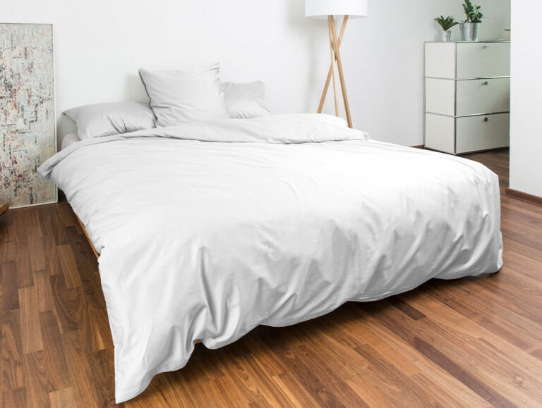 Ein ordentlich gemachtes Bett mit weißem Bettzeug steht auf einem Holzboden. Dahinter befinden sich eine Stehlampe, eine weiße Kommode mit Pflanzen darauf und eine dekorative Tafel, die an einer weißen Wand lehnt.