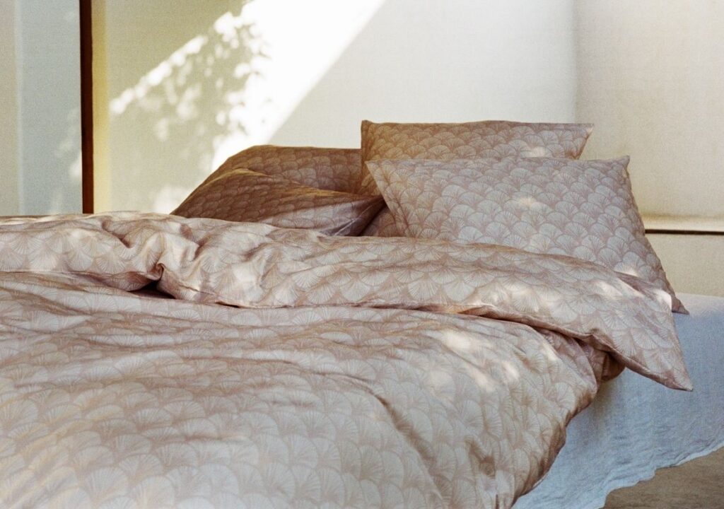 Ein ordentlich gemachtes Bettenland-Bett mit hellbeiger Bettwäsche und Kissen mit dezentem Muster, beleuchtet von sanftem natürlichem Sonnenlicht, das durch ein nahe gelegenes Fenster fällt und sanfte Schatten an der Wand erzeugt.