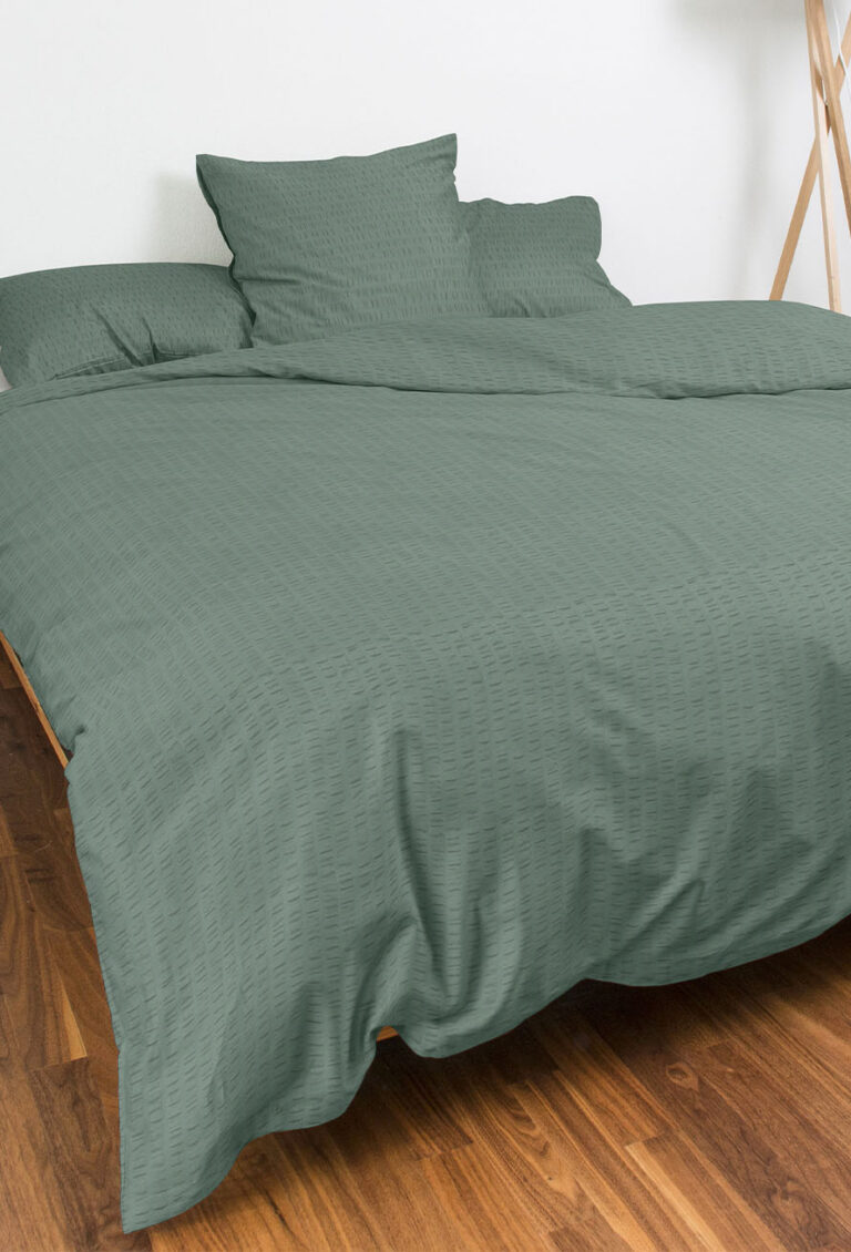 Ein ordentlich gemachtes Bett mit grüner Bettwäsche und drei passenden Kissen, auf einem Holzboden mit einer weißen Wand und einem Teil einer hölzernen Stativlampe im Hintergrund.