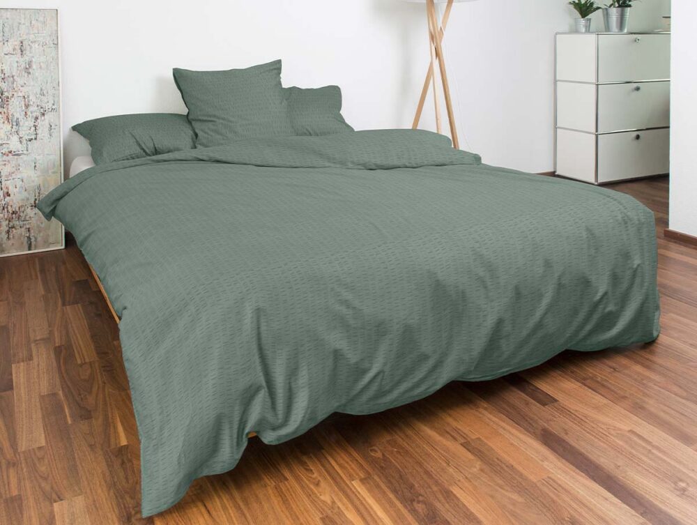 Ein ordentlich gemachtes Bett mit grünem Bettzeug und mehreren Kissen in einem modernen Schlafzimmer mit Holzfußboden, weißen Wänden, einer Stehlampe, einer weißen Kommode und einer Topfpflanze im Hintergrund.
