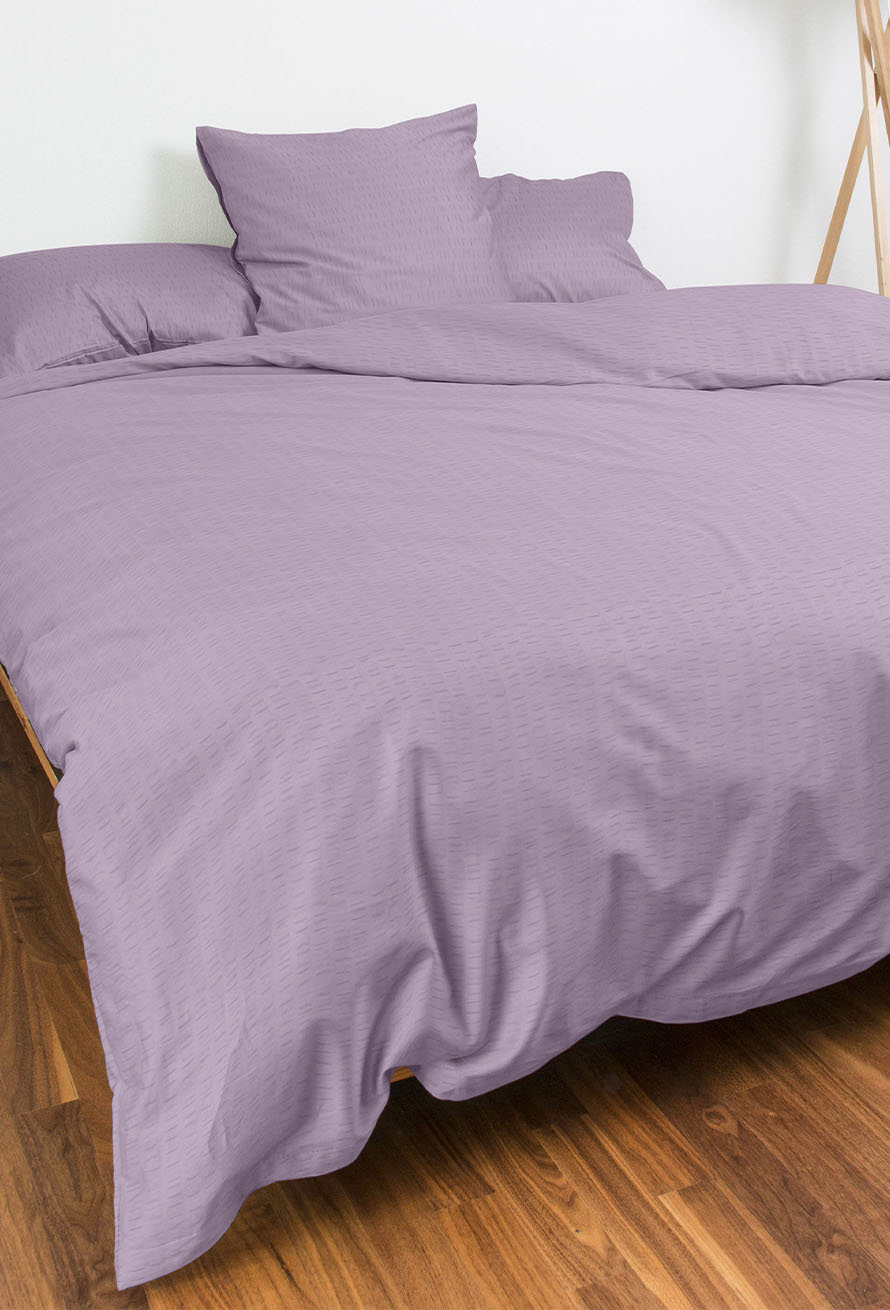 Ein ordentlich gemachtes Bett mit hellviolettem Bettzeug und zwei Kissen in einem Raum mit Holzfußboden und weißen Wänden.