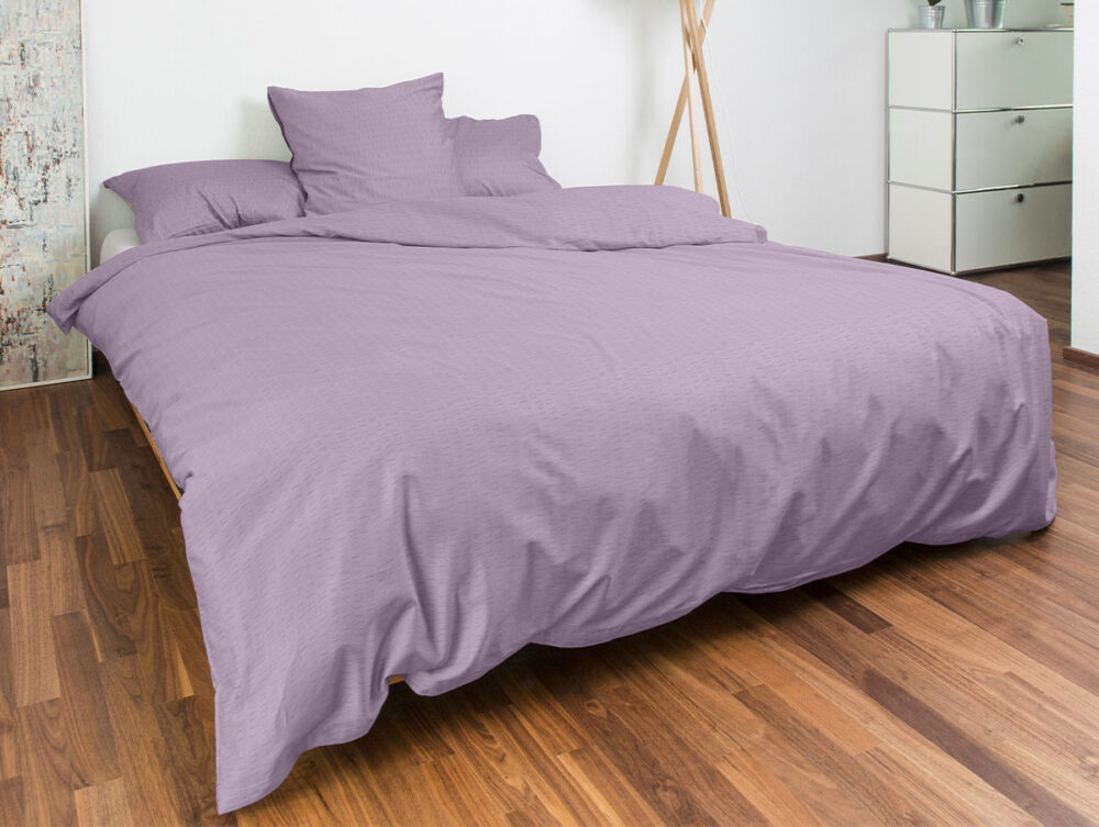 Ein ordentlich gemachtes Bett mit hellvioletter Bettwäsche und Kissen steht in einem modernen Schlafzimmer mit Holzfußboden, weißen Wänden, einer Kommode und einigen dekorativen Gegenständen im Hintergrund.