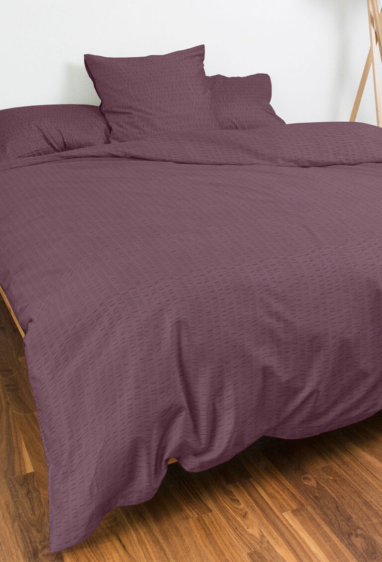 Ein ordentlich gemachtes Bett mit einem strukturierten lila Bettbezug und passenden Kissen, auf einem Holzboden in einem minimalistischen, hellen Raum.